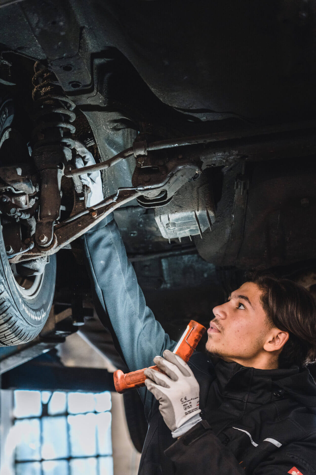Junger Mechaniker von Kayki Autoservice bei der Inspektion des Fahrwerks eines Autos, während er in der Werkstatt in Hildesheim einen Schlagschrauber hält.